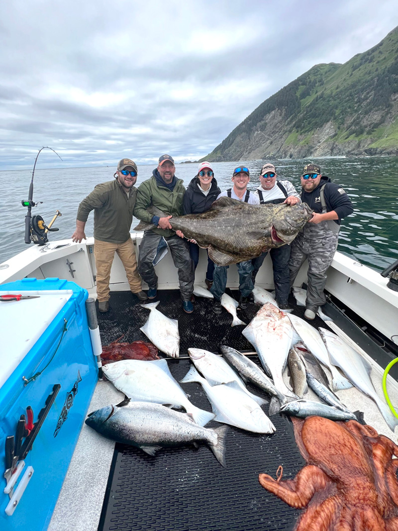 Six fishers hold up one massive sturgeon with a pile of fish and a very large octopus in front of them.