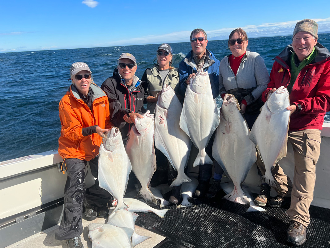 Six fishers hold up a halibut each with several halibut on the ground.